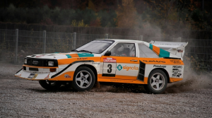 Imagem gerada por IA de um carro clássico de rali com wraps de corrida laranja, branco e turquesa a deslizar numa pista de gravilha.